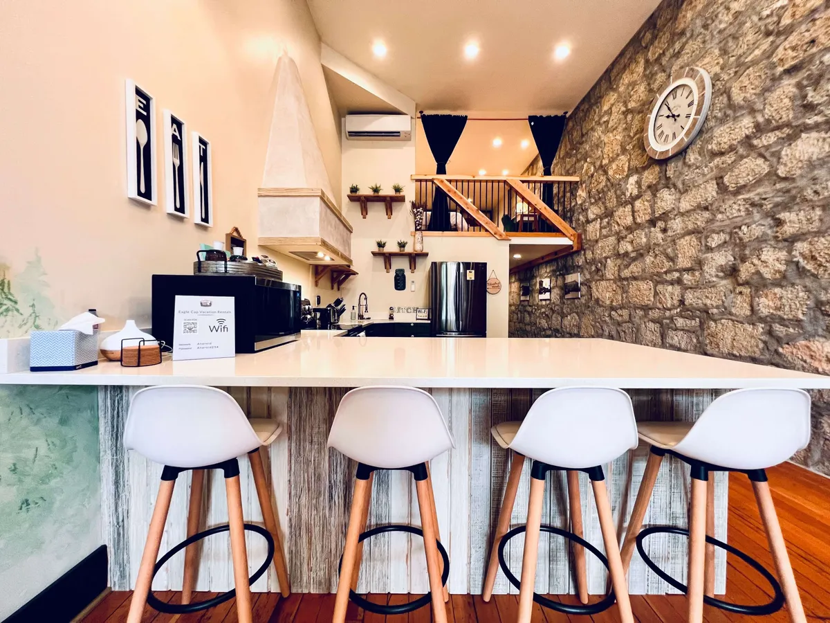 A view of a kitchen island with stools, set against a rustic stone wall with a decorative clock, and an open loft area visible above. The space is arranged for social interaction, with comfortable seating and a modern, inviting atmosphere perfect for entertaining guests or enjoying family meals.