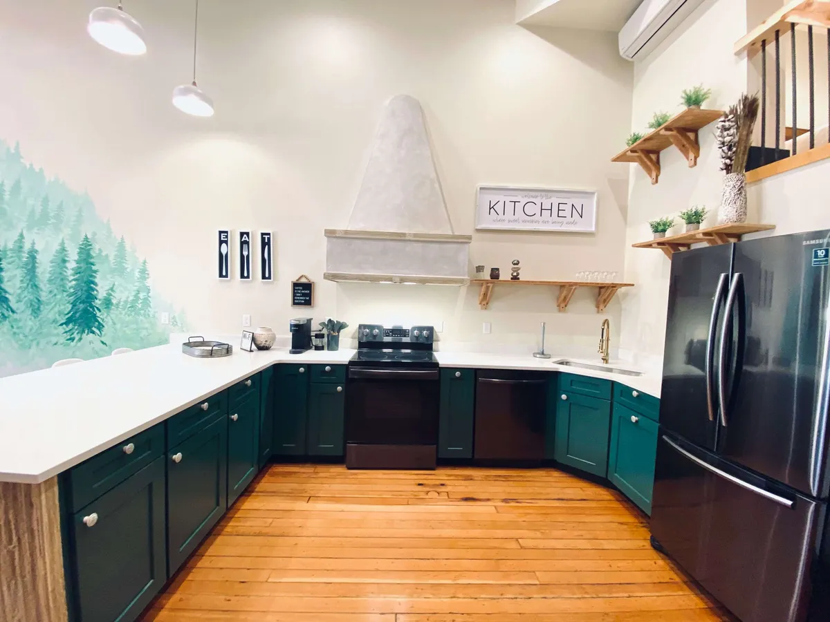 This image showcases a stylish kitchen with green cabinetry, a pristine white quartz countertop, open wooden shelves, kitchen signage, and stainless steel appliances. The overall ambiance is cozy and contemporary, with visual cues of cooking and gathering in the space.