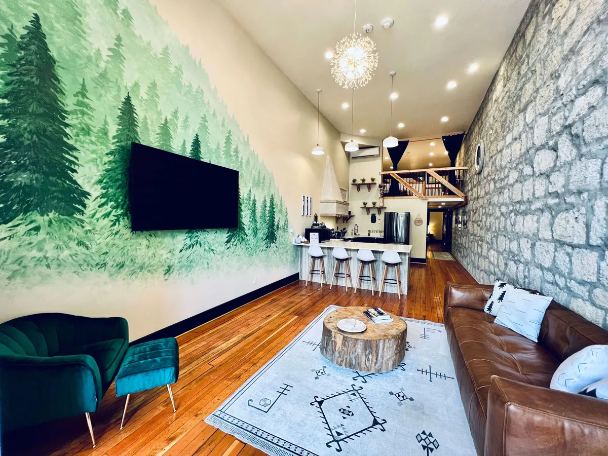 A sunlit living area with a forest mural, modern kitchen island, exposed stone wall, a wooden coffee table, and comfortable seating. Morning light pours through tall windows, illuminating the plush furniture and highlighting both the historic stonework and contemporary design elements.