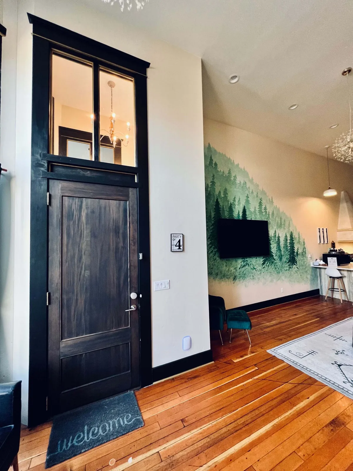The image showcases the condo's inviting entrance area, complete with a wooden door, forest mural, green chair, and a welcoming mat. The chandelier casts a warm glow, while the seating area creates an immediate sense of comfort and hospitality. The forest mural subtly connects the interior atmosphere to the natural beauty outside, reinforcing the outdoor adventure narrative.