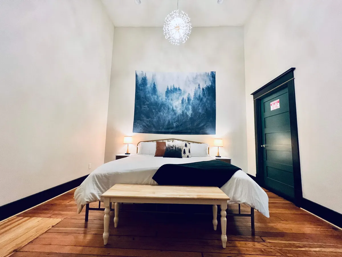 A tranquil bedroom scene with a plush bed set against a captivating forest mural, unique chandelier, and softly glowing bedside lamps. The space exudes peacefulness and creative flair, suggesting both retreat and inspiration.