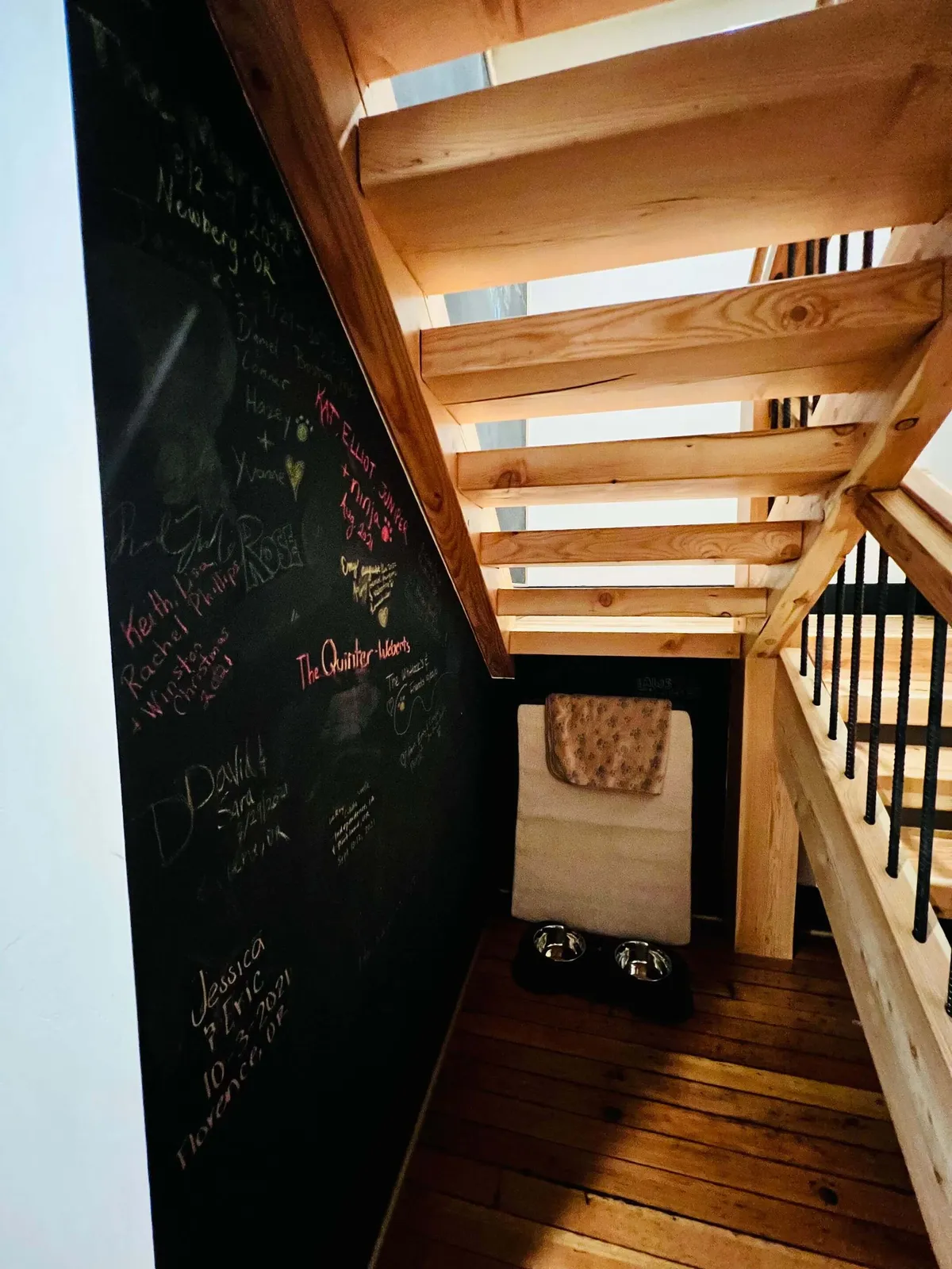 This image centers on a chalkboard wall adorned with messages, with a staircase leading to another part of the home. Notably, pet bowls and a folded blanket are visible in the foreground, suggesting the presence and comfort of pets within the space. The environment feels warm, homey, and welcoming, inviting both guests and their animals to relax.