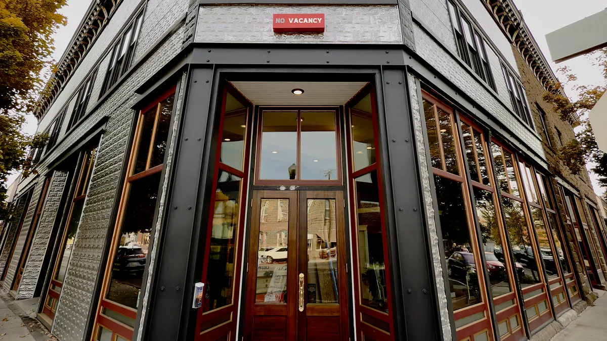 A warm and engaging exterior shot of the Litch Hotel’s entrance, featuring inviting doors, a 'NO VACANCY' sign, and large windows that showcase the building's architectural character. The bustling sidewalk and nearby shops evoke a sense of discovery and activity as guests arrive to explore the area.