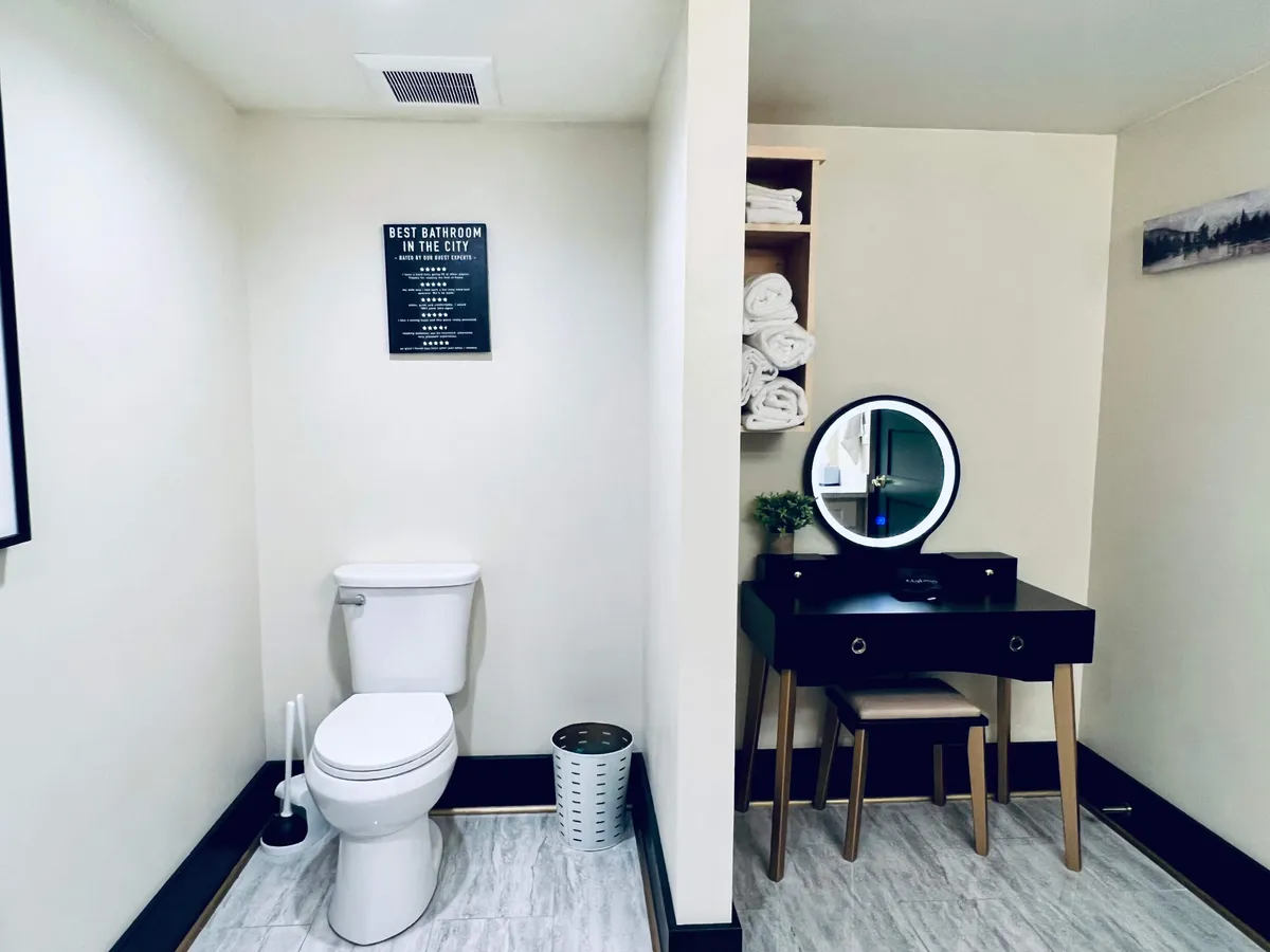 A functional and inviting bathroom scene with a vanity mirror, neatly arranged towels on a shelf, a decorative plant, and a tasteful sign on the wall. The lighting is bright, emphasizing cleanliness and comfort for guests preparing for the day or winding down.