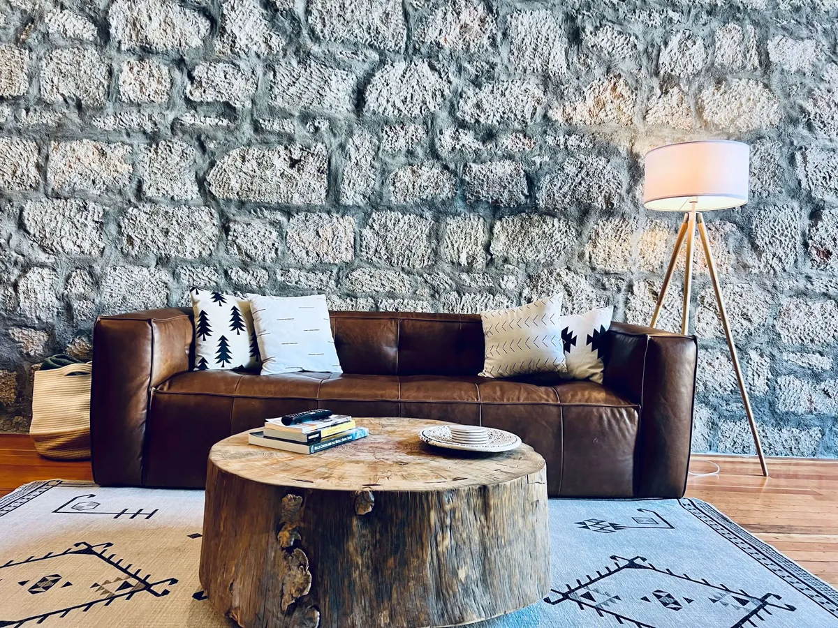 The image showcases a cozy living area featuring a rich leather sofa, a rustic wooden coffee table, a warm floor lamp, and a textured stone wall that hints at the building's historic charm. The inviting scene is softly lit, encouraging relaxation and conversation.